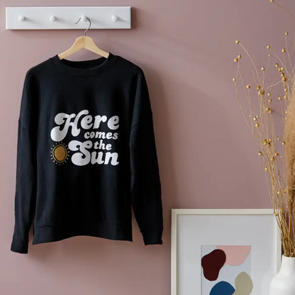 A black crewneck sweatshirt hanging on a wooden hanger against a dusty pink wall. The sweatshirt features a graphic print that reads 'Here comes the Sun' in white script, accompanied by a small gold sun illustration.