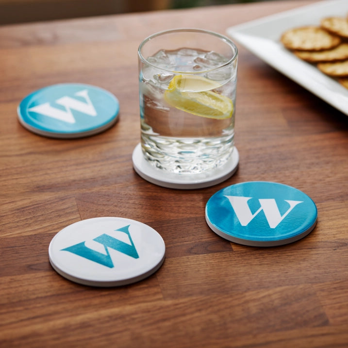Three round white coasters with blue and teal letter 'W' designs are arranged on a wooden table, with one coaster supporting a glass of water with a lemon slice and another holding a cracker in the background.