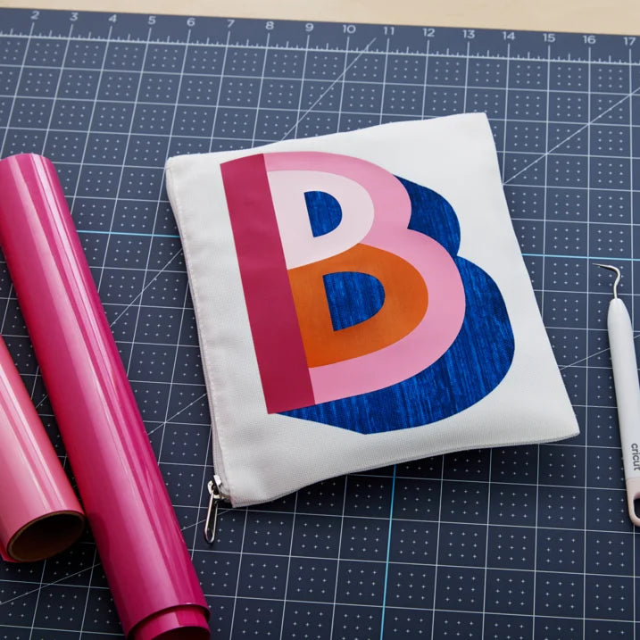 A white zippered pouch featuring a large layered letter 'B' design in pink, orange, and blue, displayed on a dark gridded cutting mat alongside craft supplies.