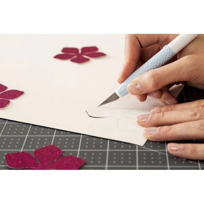 A close-up of a person's hands using a craft knife to cut out a flower shape from white cardstock, with matching magenta petal cutouts scattered on a gray gridded cutting mat.