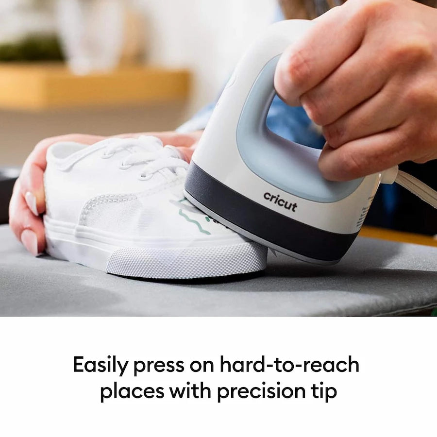 A person uses a white and gray Cricut ironing iron to press a white sneaker on a gray ironing board, with text below reading 'Easily press on hard-to-reach places with precision tip'.
