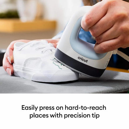 A person uses a white and gray Cricut ironing iron to press a white sneaker on a gray ironing board, with text below reading 'Easily press on hard-to-reach places with precision tip'.