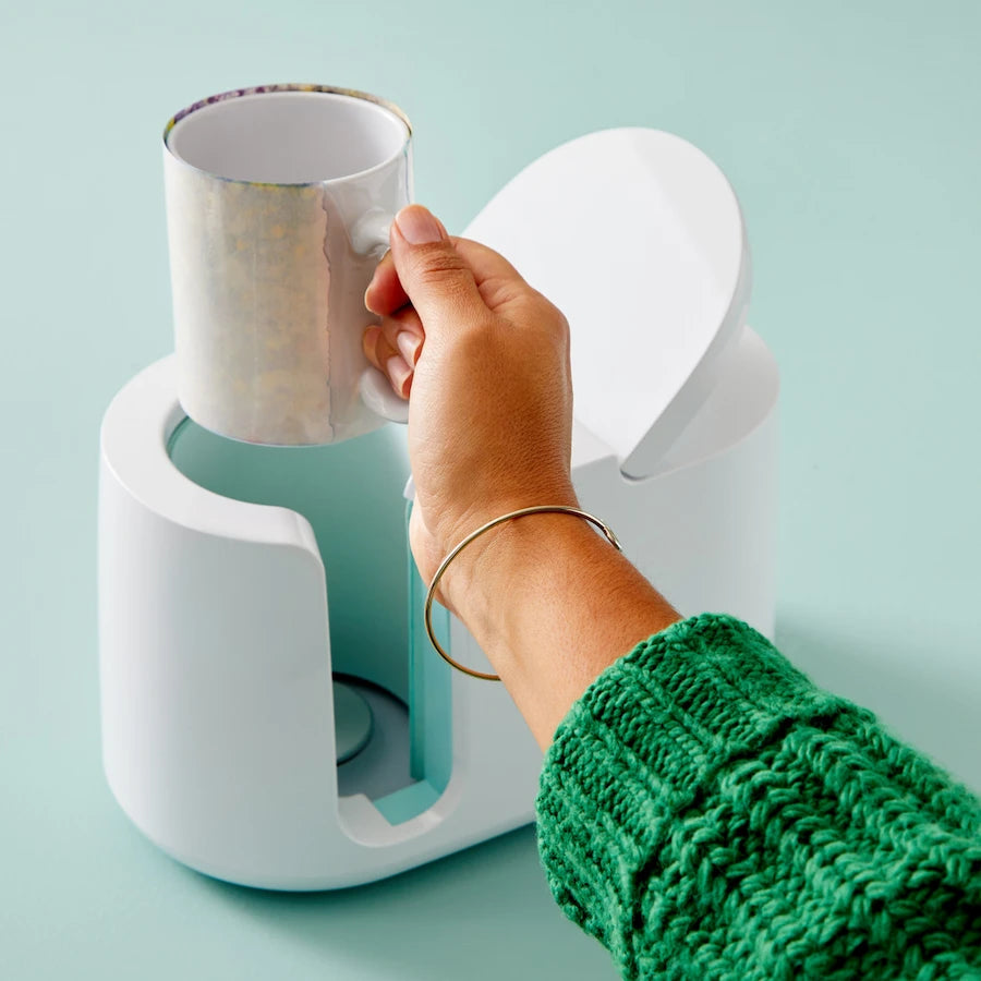 A hand wearing a green knit sweater and a gold bracelet holds a metallic silver mug over a white and light blue coffee maker on a light blue background.