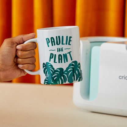 Hand holding a mug with 'Paulie the Plant' design in front of a Cricut machine.
