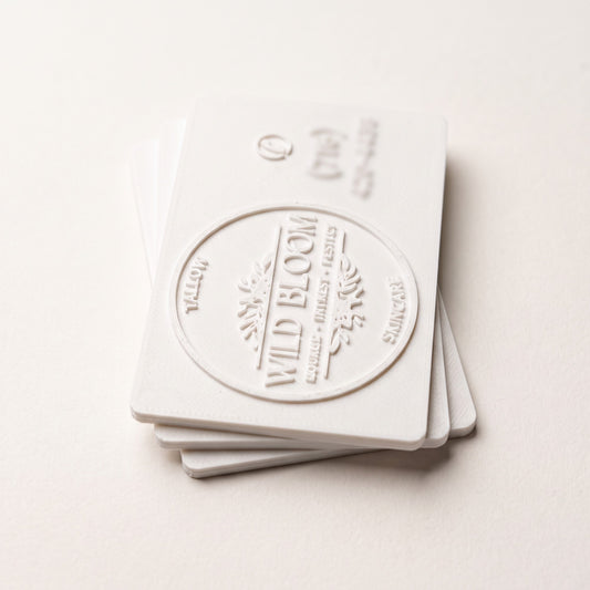 Stack of white 3D printed business cards with a raised relief logo, side-lit to show the layered texture and card edge