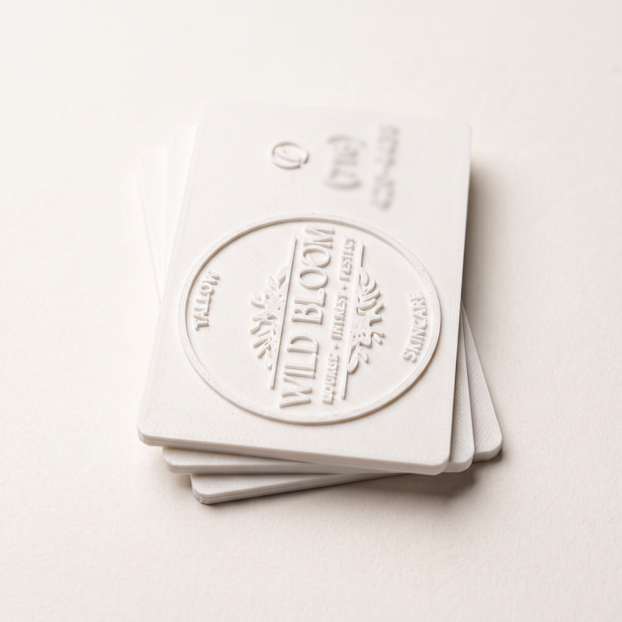 Stack of white 3D printed business cards with a raised relief logo, side-lit to show the layered texture and card edge