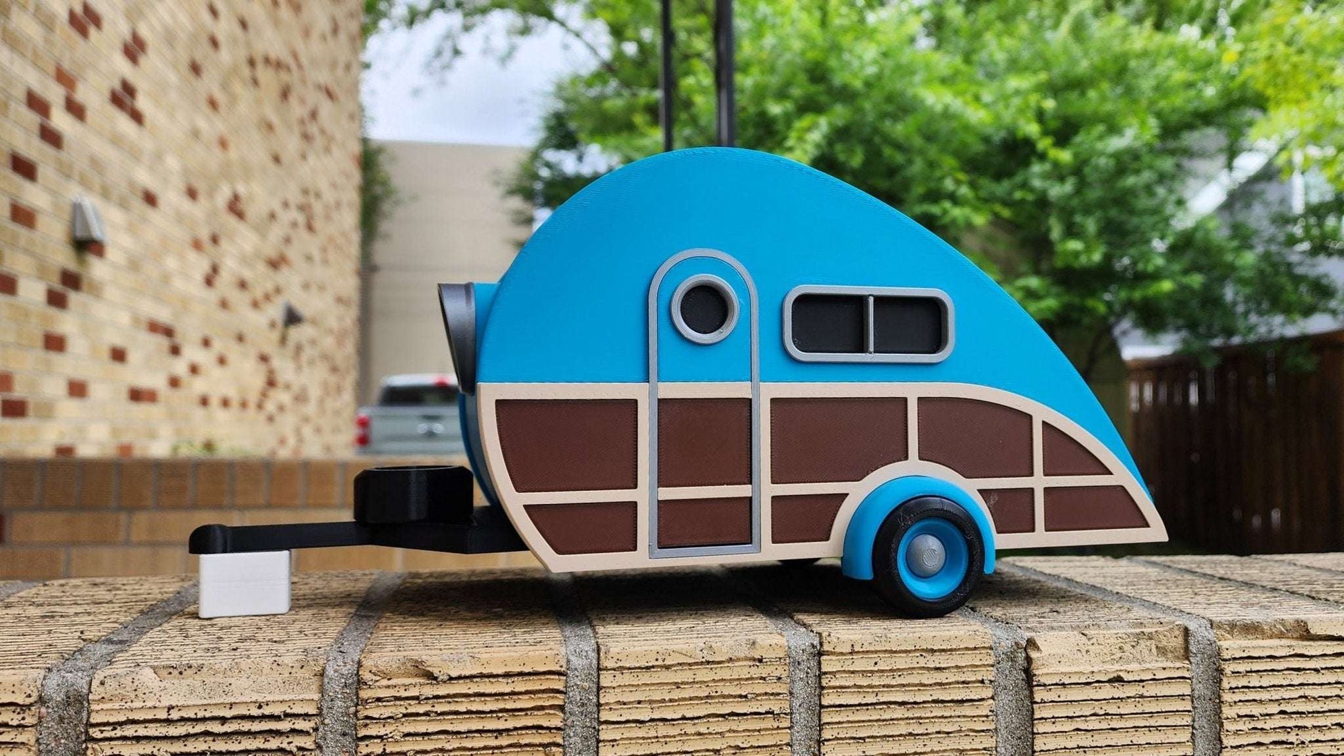 Model of a blue and brown camper on a wooden platform with trees and buildings in the background.
