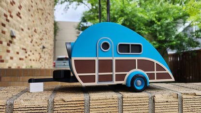 Model of a blue and brown camper on a wooden platform with trees and buildings in the background.